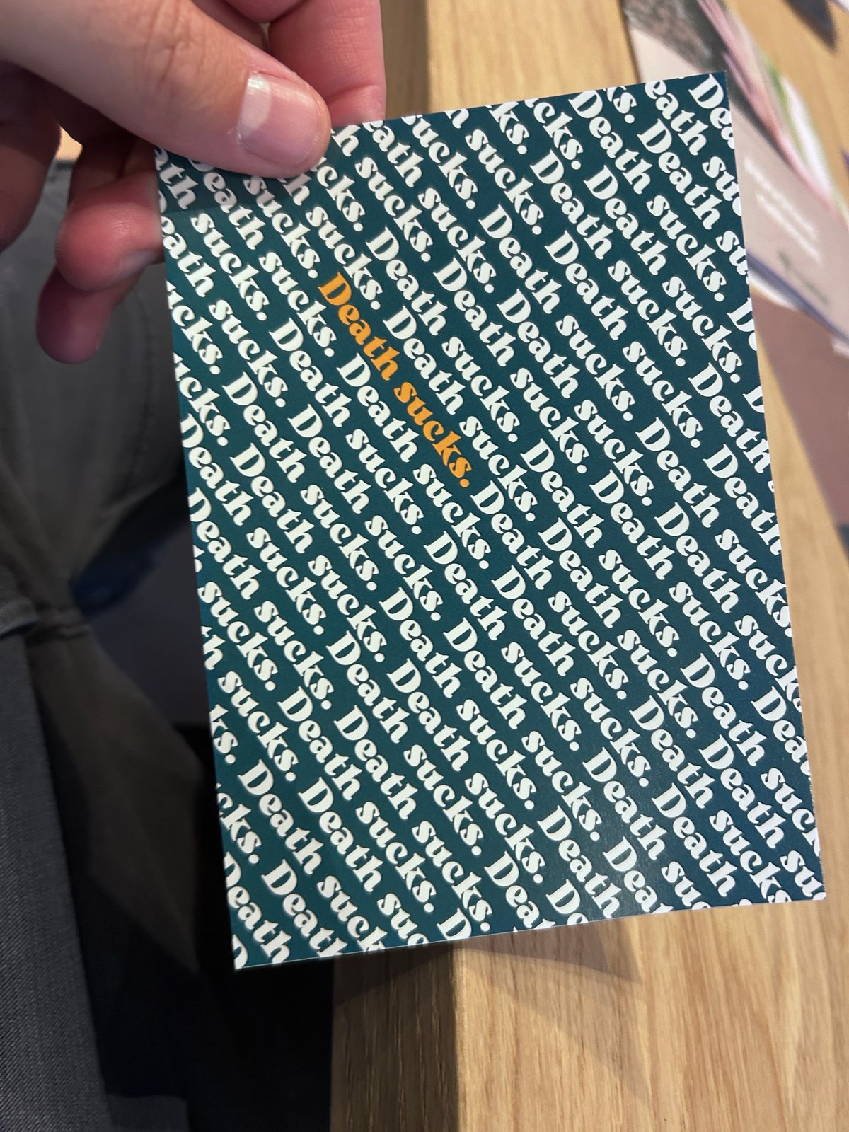 Postkarte, auf der der Satz „Death sucks.“ dicht an dicht in Weiß auf Dunkelgrün steht, einmal davon in Orange hervorgehoben