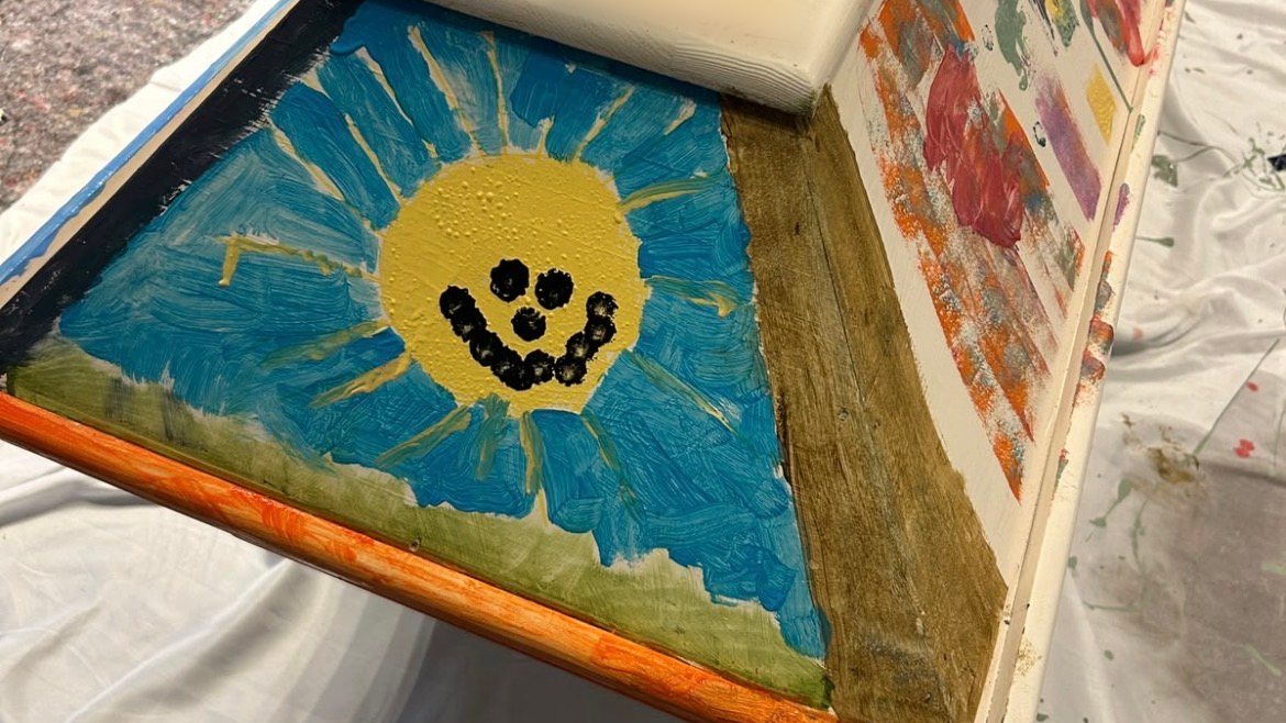 Von Kindern bemalte Stirnseite eines Sargs: eine gelbe Sonne mit lachendem Gesicht auf blauem Himmel über einer grünen Wiese